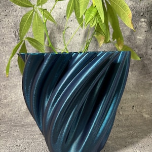 Op de afbeelding: Een tealblauwe, 3D-geprinte plantenpot met een draaiend, textuurontwerp. Een groene plant met grote bladeren groeit in de pot.
