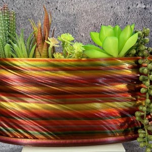 Op de afbeelding: Een decoratieve, ovale plantenbak met een wervelend patroon van rood, goud en groen. De plantenbak is gevuld met diverse kunstmatige vetplanten en planten, waaronder cactussen en parelsnoer. De plantenbak staat op een bijpassende schotel. Lengte: circa 30 cm.