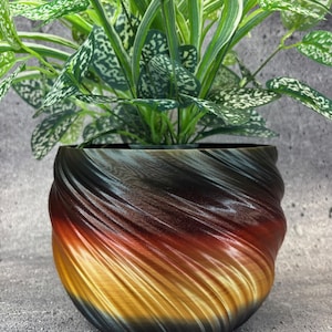 May include: A decorative plant pot with a swirling design in shades of black, brown, red, and gold. The pot contains a green plant with white speckled leaves. The pot is sitting on a gray surface.