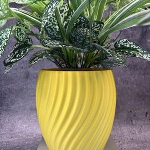 May include: A bright yellow plant pot with a vertical spiral ribbed texture. The pot contains a lush artificial plant featuring variegated green and white leaves with speckled patterns, creating a dense foliage display above the rounded container.