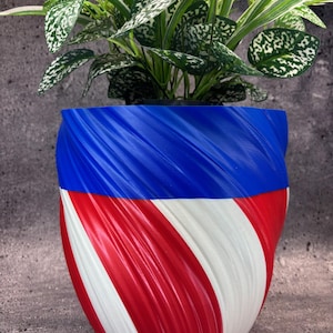 May include: A decorative plant pot with a spiral design in red, white, and blue. The top section is blue, transitioning to red and white stripes. The pot holds a green plant with white speckled leaves, adding a natural element to the design.