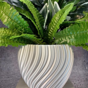Op de afbeelding: Een decoratieve plantenarrangement met levendig groen blad in een witte, swirl-patroon keramische pot. De pot staat op een lichtgekleurde, rechthoekige basis, wat zorgt voor een moderne en stijlvolle presentatie.