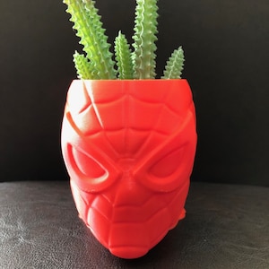 Puede incluir: Una maceta roja impresa en 3D con forma de máscara de superhéroe. La maceta está llena de una pequeña planta suculenta verde.