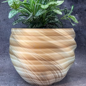 Pode incluir: Um vaso decorativo, castanho claro e branco, com redemoinhos, contendo uma planta verde e branca manchada. O vaso tem um design único e texturizado com uma forma arredondada. A planta tem folhas longas e finas com manchas brancas e verdes.