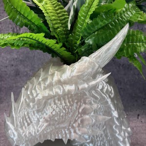 May include: A pearlescent white dragon head planter with a detailed, textured surface. The planter is filled with vibrant green artificial plants. The dragon head has sharp horns and a detailed snout. The planter sits on a light gray surface.