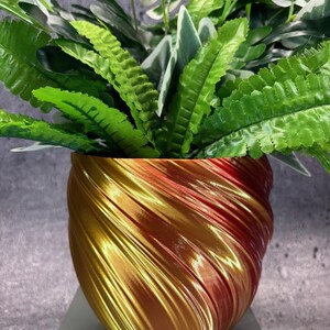May include: A decorative gold and red-toned planter with a swirling design, filled with vibrant green and white artificial plants. The planter sits on a white surface, creating a striking contrast. The overall aesthetic is modern and stylish.