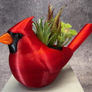 Puede incluir: Una maceta en forma de cardenal rojo brillante, con pico naranja y detalles negros. La maceta está llena de una variedad de suculentas artificiales verdes y marrones. Esta pieza decorativa añade un toque de naturaleza a cualquier espacio.