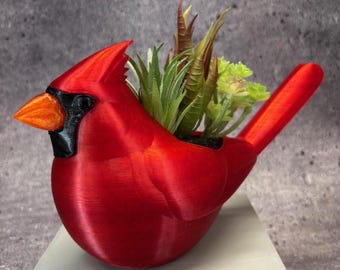 Puede incluir: Una maceta en forma de cardenal rojo brillante, con pico naranja y detalles negros. La maceta está llena de una variedad de suculentas artificiales verdes y marrones. Esta pieza decorativa añade un toque de naturaleza a cualquier espacio.