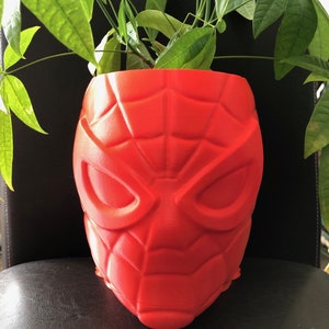 Puede incluir: Una maceta de plástico roja con forma de cabeza de Spider-Man con una planta verde en su interior.