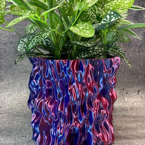 May include: A colourful, abstract-patterned plant pot in blue, purple, and red, containing a vibrant green plant with speckled leaves. The textured surface of the pot adds visual interest, creating a modern and artistic aesthetic. The plant pot is a decorative item.