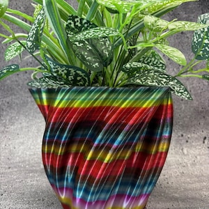 May include: A colorful, striped, 3D printed planter with a green plant with white speckles. The planter has a swirling, abstract design with red, yellow, blue, green, and purple stripes.