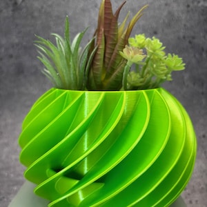 May include: A bright green, 3D printed planter with a swirling design. The planter is filled with a variety of succulents, including a green air plant and a red-brown succulent.