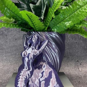 May include: A purple mermaid planter with a detailed design, holding her arms above her head. The planter is filled with green artificial plants. The mermaid's tail and hair are flowing. The planter is on a gray surface.