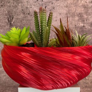May include: A vibrant red, textured planter filled with a variety of succulents and cacti. The planter has a unique, flowing design. The plants include a bright green succulent, a tall cactus, and other small, colorful plants. The planter is sitting on a white surface.