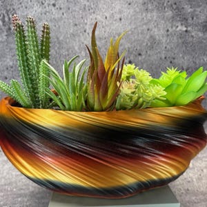 May include: A decorative planter with a swirling design in shades of black, orange, and gold, filled with a variety of artificial succulents and cacti. The planter has a unique, textured appearance, adding a touch of modern style to any space.