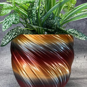 May include: A plant pot with a spiraling, ribbed texture featuring a gradient of metallic copper, bronze, and dark brown tones. The pot contains a lush, green plant with variegated leaves that have white and light green speckled patterns.