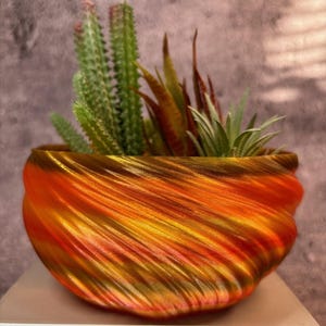 Puede incluir: Un macetero de espiral impreso en 3D de color naranja, amarillo y dorado con una variedad de suculentas que crecen en su interior.