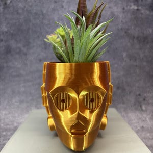 May include: A metallic gold-colored planter shaped like a robotic head with circular eyes and geometric side details. The planter contains a variety of green and brownish-purple artificial succulent plants extending from the top opening.