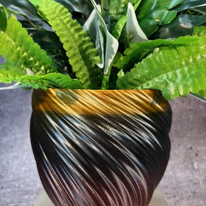Op de afbeelding: Een zwarte en gouden swirl-pot met een groene, bladerrijke plant erin. De pot heeft een glanzende, metallic afwerking.