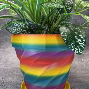 Puede incluir: Una maceta geométrica de colores arcoíris con un platillo amarillo. La maceta está hecha de plástico impreso en 3D y tiene un diseño único y moderno. La maceta está llena de una planta verde con manchas blancas.