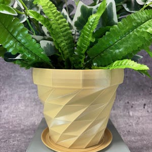 May include: A light yellow, geometric-patterned flower pot with a matching saucer holds a variety of green plants. The pot has a textured, diamond-like design. The plants include ferns and ivy.