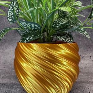 May include: A gold, swirl-patterned planter with a green leafy plant inside. The planter is made of a shiny, metallic material.