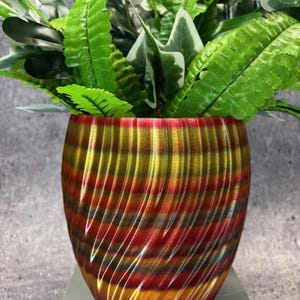 May include: A decorative, multi-colored plant pot with a swirling design in shades of red, gold, and brown. The pot is filled with a variety of green plants with different leaf shapes and textures. The pot sits on a white surface.