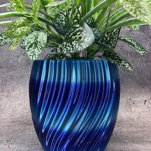 Puede incluir: Una vibrante maceta azul, con textura y patrón en espiral, que contiene una exuberante planta verde con hojas moteadas de blanco. El diseño de la maceta presenta ranuras verticales que crean un efecto visual dinámico. La planta tiene hojas largas y delgadas con rayas blancas.