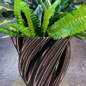Op de afbeelding: Een zwarte, 3D-geprinte plantenpot met een draaiend, textuurontwerp. De pot is gevuld met groene en witte bladplanten.