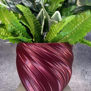 May include: A burgundy-coloured, spiral-textured planter pot filled with various green plants. The plants have different shades of green, with some leaves featuring white edges. The pot sits on a light-coloured surface, creating a contrast with the dark pot.
