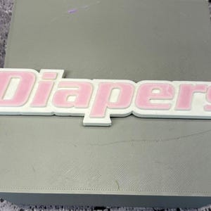 Puede incluir: Un letrero blanco con la palabra "Diapers" en rosa, colocado sobre una superficie gris. El letrero tiene un efecto tridimensional en relieve, con las letras rosas ligeramente empotradas dentro del contorno blanco.