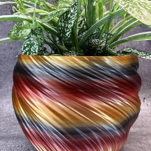 May include: A round planter with a spiralling, ribbed texture featuring a gradient of metallic colours including copper, bronze, black, and deep red. A green and white variegated houseplant with pointed leaves grows out of the top of the planter.