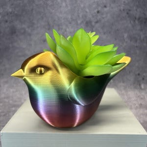 Op de afbeelding: Een vogelvormige plantenbak met een gladde, verloopafwerking die overgaat van goud naar groen, blauw en paars. De plantenbak bevat een felgroene kunstmatige vetplant met gelaagde, puntige bladeren, wat zorgt voor een levendige en kleurrijke uitstraling.