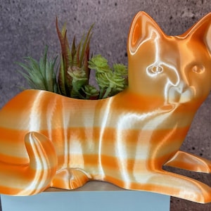 May include: A 3D-printed orange and white striped cat planter holding small green plants. The cat is in a reclining position with its paws extended. The planter is made of plastic and has a glossy finish.