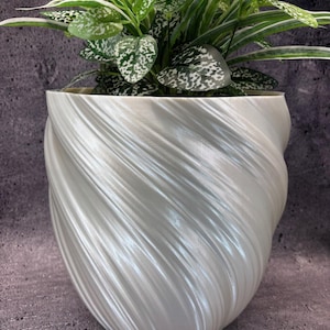 Puede incluir: Una maceta blanca con textura en espiral que contiene una planta de interior con hojas verdes y blancas moteadas. La maceta tiene un diseño único y arremolinado y está llena de exuberante vegetación.