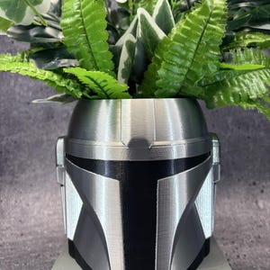 May include: A 3D-printed planter shaped like a silver and black helmet with a T-shaped visor. The helmet features a metallic silver finish on the upper dome and side panels, contrasted by a matte black central faceplate. Green artificial foliage is placed inside.