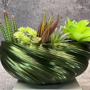 Puede incluir: Una maceta de color verde oscuro metalizado con una textura acanalada en espiral que contiene un surtido de suculentas artificiales. El arreglo incluye cactus verdes espinosos, plantas de hojas verde claro, follaje puntiagudo marrón rojizo y una gran roseta verde brillante.