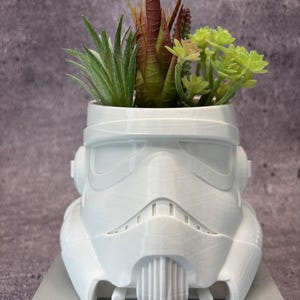 Peut inclure: Pot de fleurs blanc en forme de casque de Stormtrooper, rempli de succulentes variées. Le pot est imprimé en 3D et repose sur une base rectangulaire grise. Les succulentes sont vertes et brun rougeâtre.