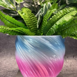 May include: A decorative planter with a gradient color scheme of pink, white, and blue. The planter has a spiral design and is filled with green and white leafy plants. The pot is sitting on a light-colored surface.