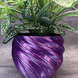 Op de afbeelding: Een glanzende, dieppaarse plantenpot met een opvallende, spiraalvormige textuur. De pot heeft een ronde, bolvormige vorm en bevat een weelderige kunstplant met bonte groene en witte bladeren die vanuit het midden naar buiten waaieren.