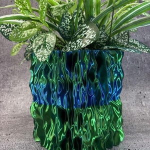 Puede incluir: Una maceta decorativa verde y azul iridiscente con un diseño ondulado texturizado. La maceta contiene una variedad de plantas verdes con hojas moteadas blancas. El diseño único de la maceta añade un toque moderno a la decoración del hogar.