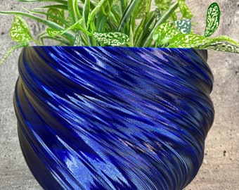 Könnte beinhalten: Ein leuchtend blauer Blumentopf mit einem strukturierten Wirbelmuster, gefüllt mit einer grünen Pflanze mit weiß gefleckten Blättern. Der Topf hat eine glänzende Oberfläche und steht auf einer grauen, strukturierten Oberfläche. Die Blätter der Pflanze weisen grüne und weiße Muster auf.