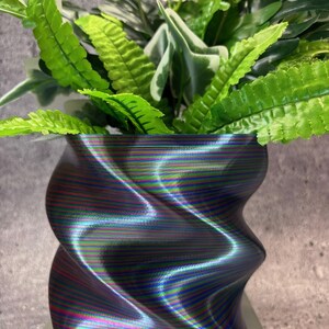 May include: A decorative, wavy-shaped planter with a multi-colored, iridescent finish. The planter holds a variety of green plants with textured leaves. The planter is sitting on a white surface. The overall theme is home decor.