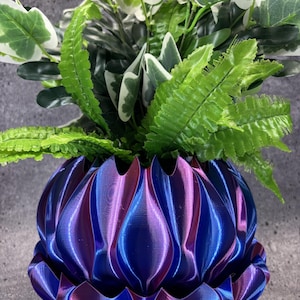 May include: A 3D printed planter with a deep, iridescent finish that shifts between metallic blue and purple tones. The vessel features a repeating, vertical, petal-like fluted design. A lush arrangement of green and white variegated foliage sits inside.