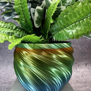 May include: A decorative planter with a swirling design in shades of green, gold, and blue, holding artificial greenery. The planter's unique texture and color gradient create a modern aesthetic. The faux plants include ferns and other leafy varieties.