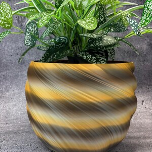 May include: A decorative plant pot with a swirling design in shades of brown, yellow, and grey. The pot holds a green plant with white-speckled leaves. The pot has a unique, textured appearance and sits on a grey surface.