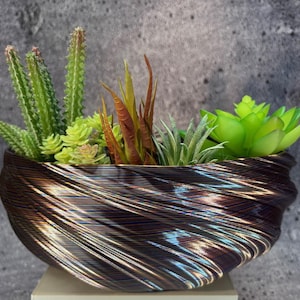 Pode incluir: Um vaso decorativo castanho e bronze preenchido com várias suculentas e cactos artificiais. O vaso tem um design único e giratório. O arranjo inclui plantas verdes, amarelas e castanho-avermelhadas.