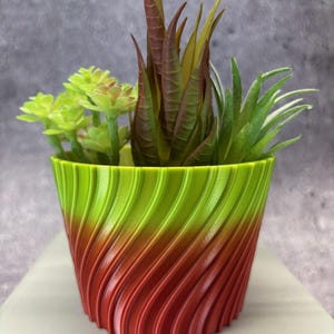 May include: A decorative planter with a spiral design, featuring a gradient of green, orange, and red. The planter holds a variety of artificial succulents in shades of green and brown, creating a vibrant display. The pot is approximately 13 cm tall.