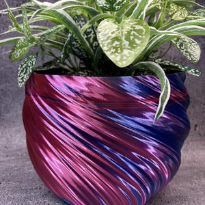 May include: A round plant pot with a metallic, iridescent finish that shifts between shades of magenta, purple, and blue. The pot features a spiraling, ribbed texture along its exterior and contains a leafy plant with variegated green and white foliage.