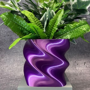 May include: A vibrant purple, wave-patterned planter holding a variety of green and white leafy plants. The planter has a glossy finish and sits on a light gray rectangular base. The plants have a mix of textures and shades of green.